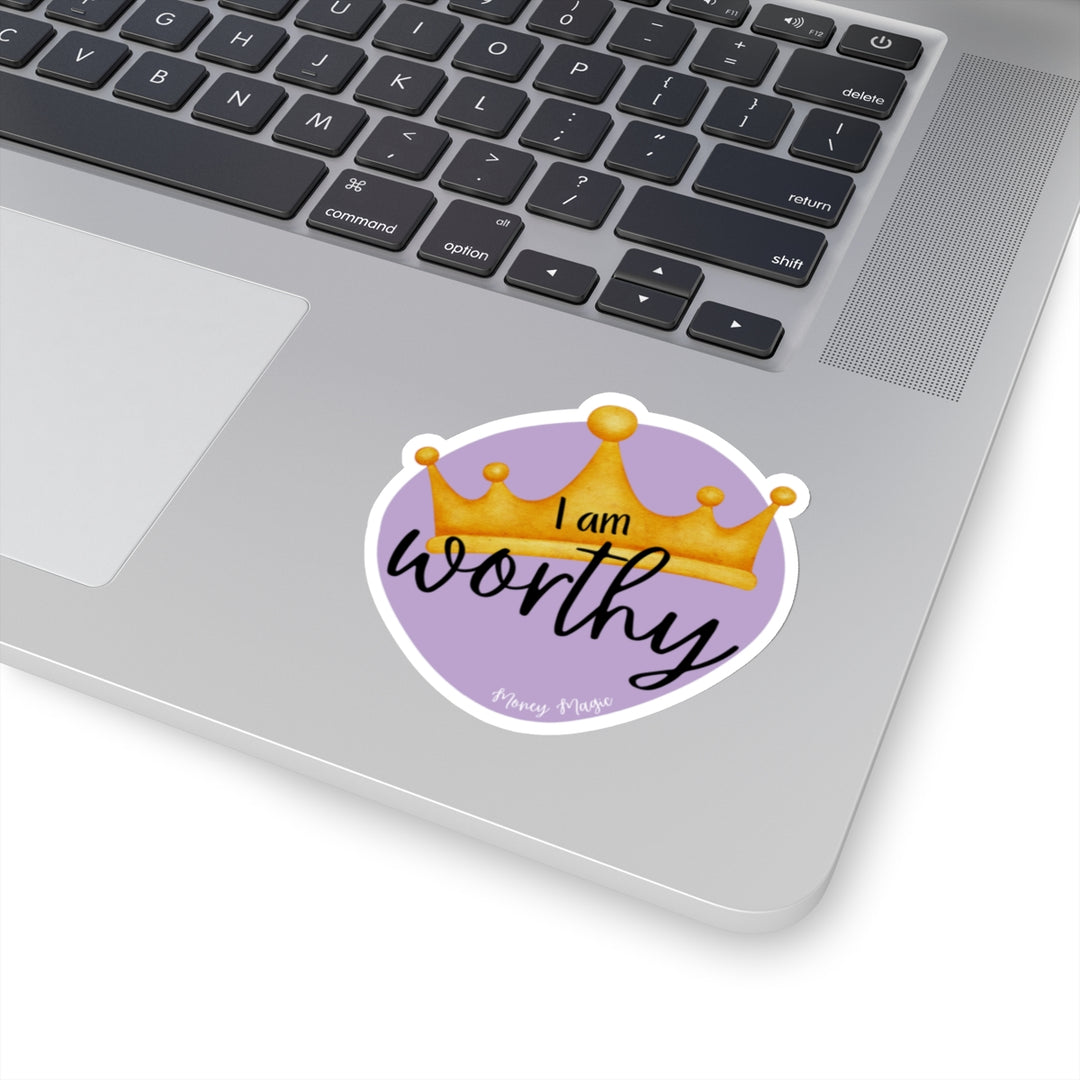 Shows a purple irregular circle with a crown in the middle. Says "I am worthy" and at the bottom "Money magic" smaller. On white background on laptop keyboard