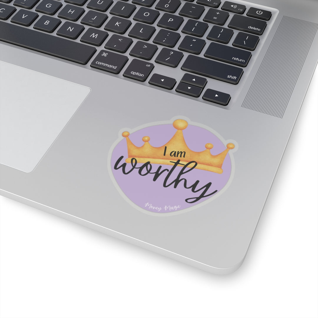 Shows a purple irregular circle with a crown in the middle. Says "I am worthy" and at the bottom "Money magic" smaller. On clear background on a laptop keyboard